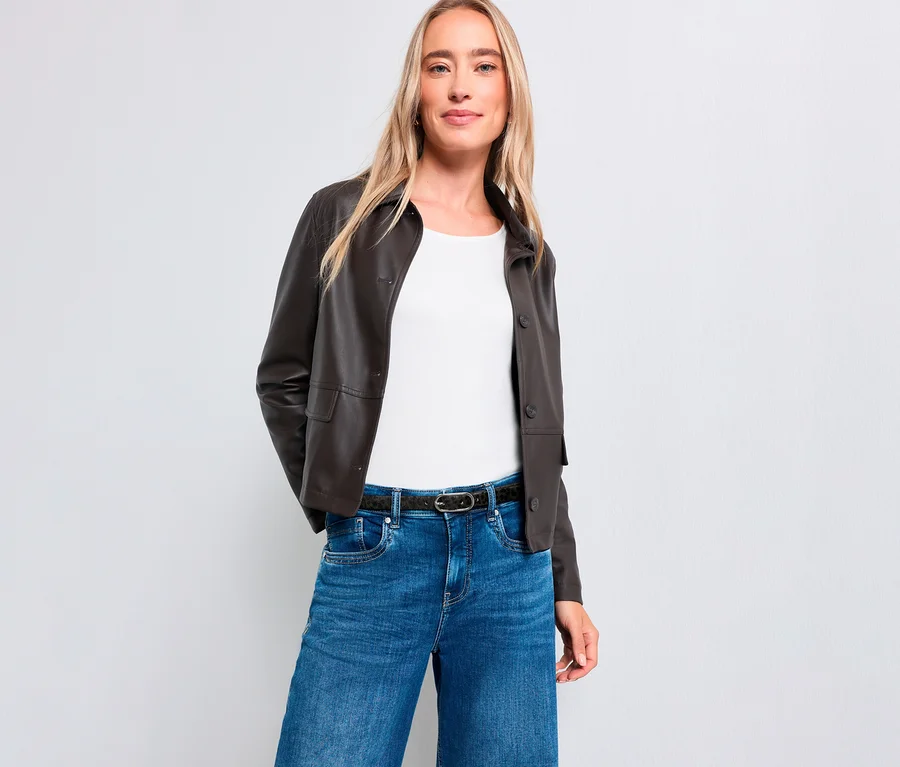 Eine blonde Frau steht lächelnd vor einem weißen Hintergrund. Sie trägt eine dunkelbraune Jacke, ein weißes Oberteil, blaue Jeans und einen dunklen Gürtel.