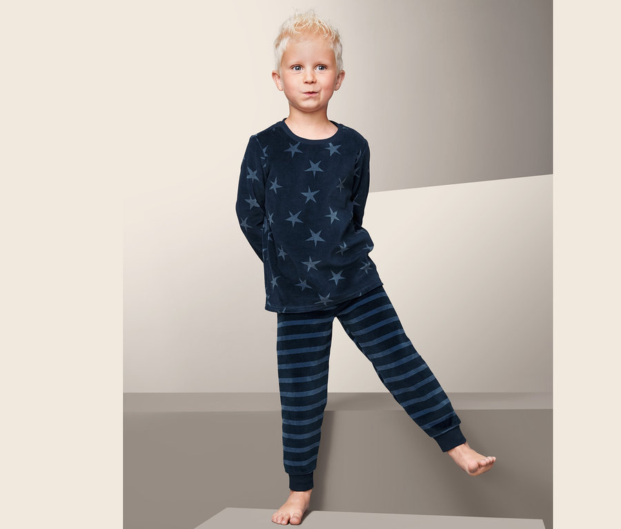 Ein Junge steht barfuß auf einem grauen Würfel und trägt einen Nicki-Pyjama mit Sternen und Streifen.
