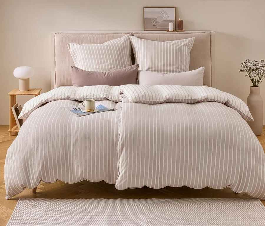 Ein Bett mit beiger und weiß gestreifter Flanell-Bettwäsche in Doppelgröße, darauf ein Magazin und eine Tasse. Links ein Nachttisch mit Lampe, rechts eine Vase mit Trockenblumen.