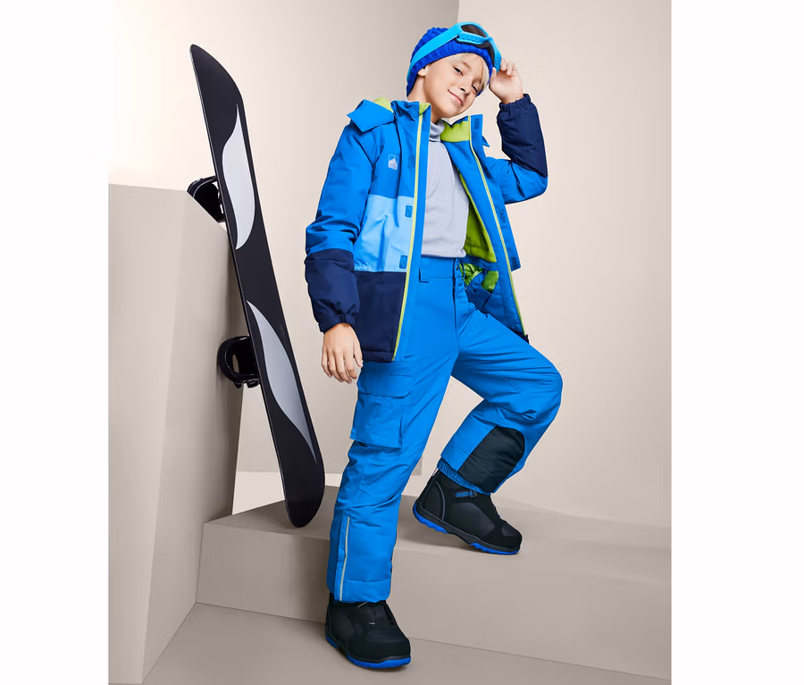 Ein Junge in einer blauen Snowboardhose steht neben einem Snowboard.