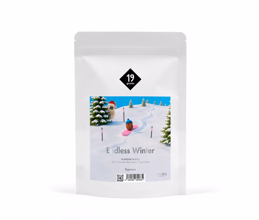 Weißer Kaffeebeutel mit Etikett, das eine Winterlandschaft zeigt: eine Kaffeebohne auf einem pinken Snowboard, eine pelzige Figur mit Weihnachtsmütze und Tannenbäume auf verschneiten Hügeln. Text: „19 grams“, „Endless Winter“, „Espresso“ und Geschmacksnoten.