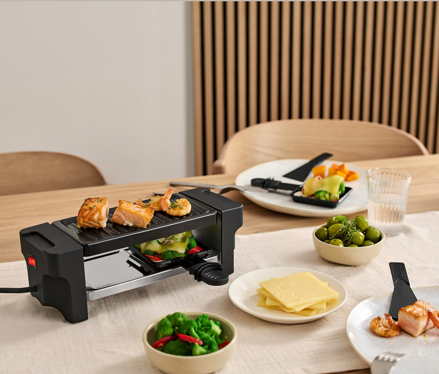 Raclette für 2 steht auf einem Tisch, mit Lachs und Garnelen auf dem Grill. Teller mit Zutaten stehen um den Grill herum.