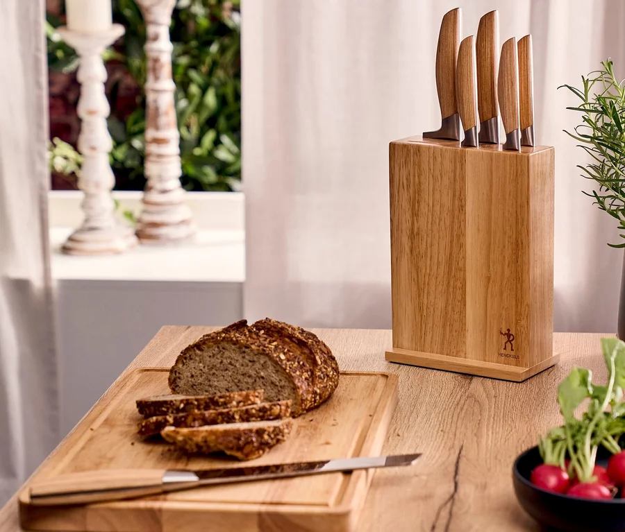Ein Henckels by ZWILLING »Pakka« Messerblockset mit fünf Messern mit Holzgriffen steht auf einer Holztheke. Daneben liegen ein angeschnittenes Brot auf einem Schneidebrett und eine Schale mit Radieschen.