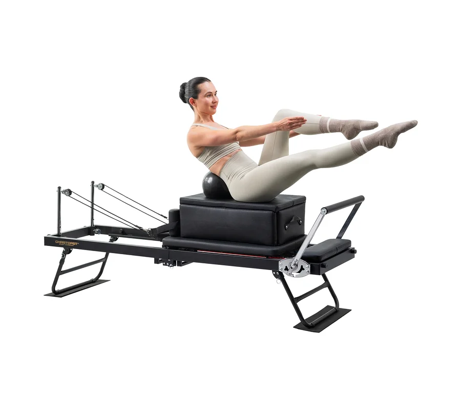Eine Frau in heller Sportkleidung trainiert auf einem schwarzen Pilates-Reformer, balancierend auf einem schwarzen Ball mit angehobenen Beinen und ausgestreckten Armen.