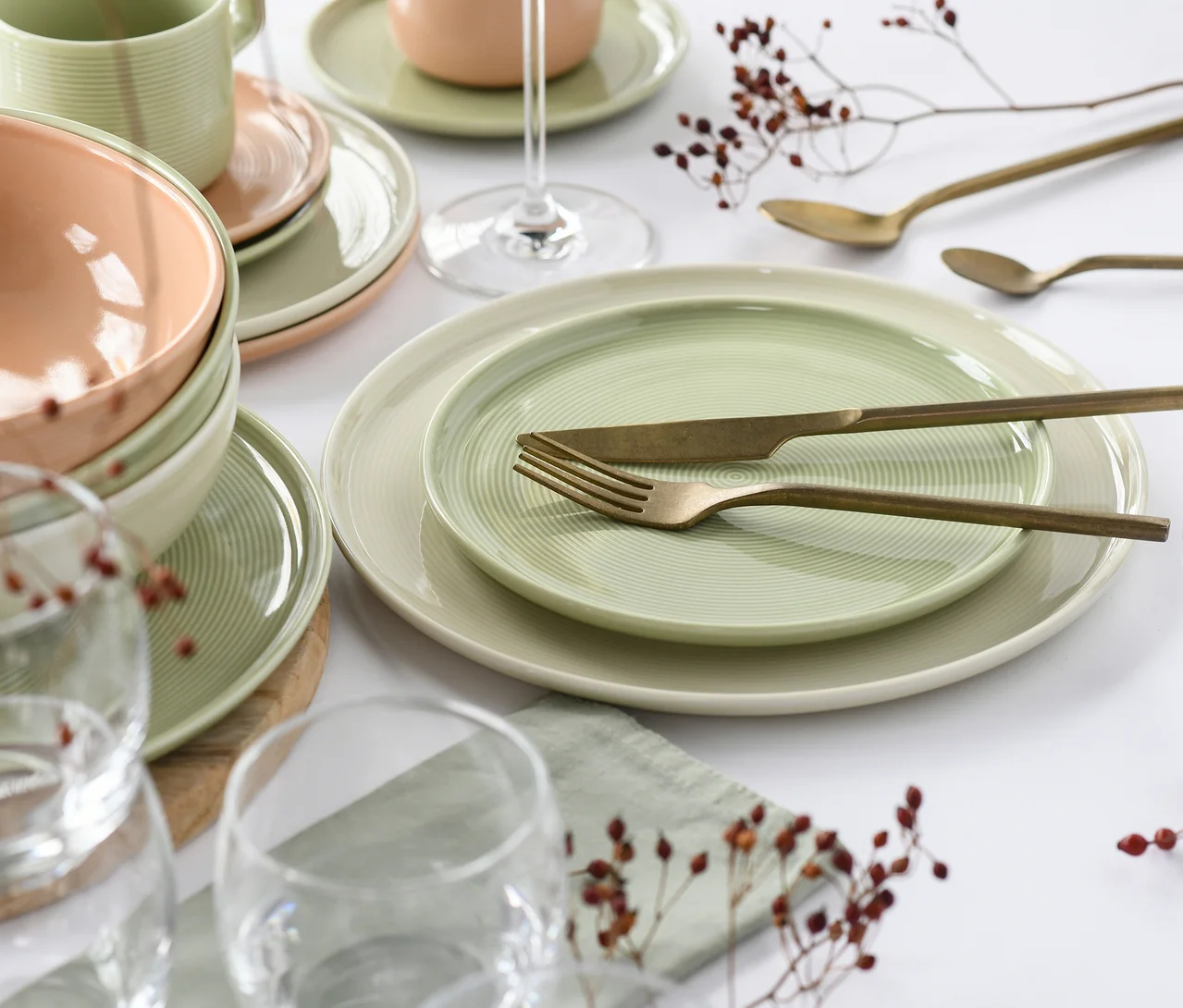 Ein creaTable Service »Session Romance« Geschirrset mit hellgrünen und pfirsichfarbenen Tellern, Schalen und einem Becher, auf einem weißen Tisch mit goldenem Besteck, Weingläsern und getrockneten Beeren.