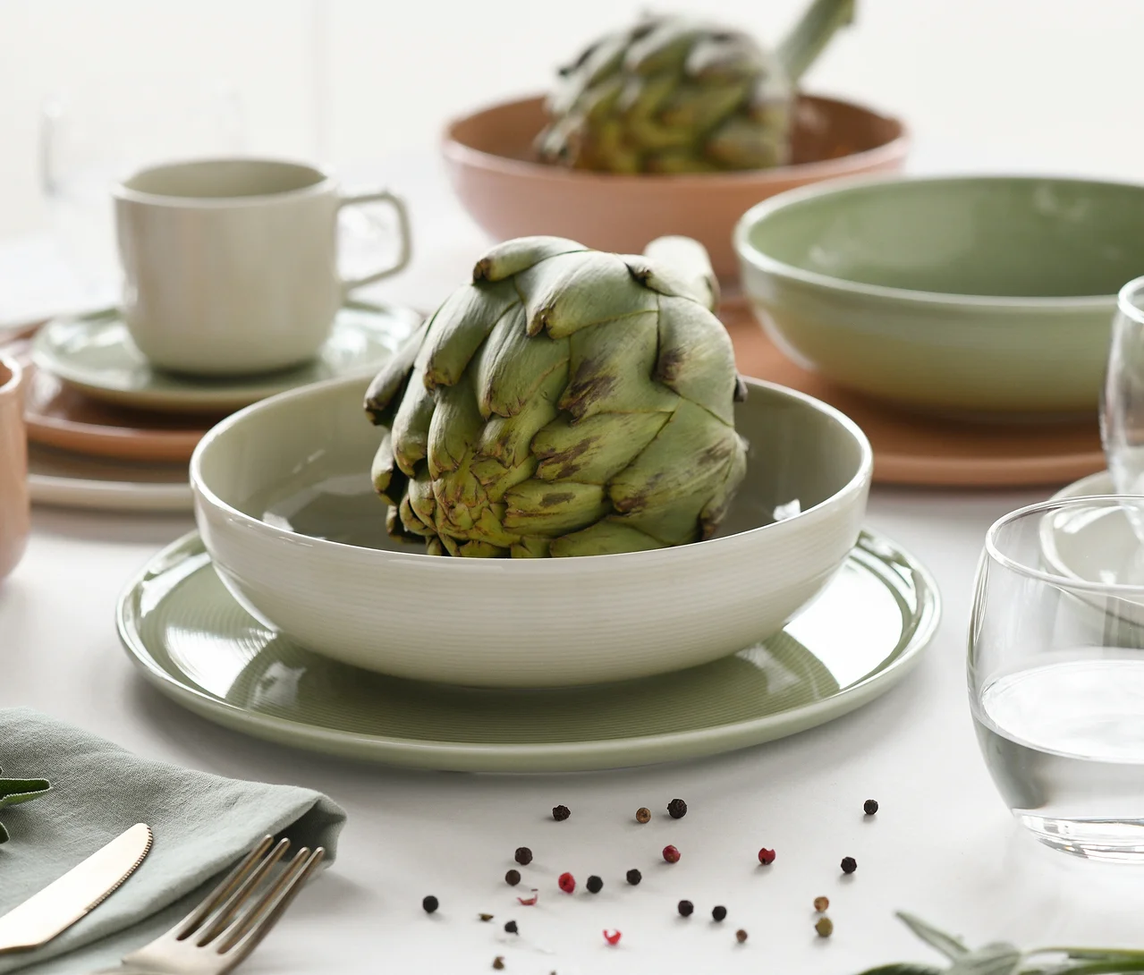 Ein Tisch gedeckt mit creaTable Service »Session Romance« Geschirr in Grün- und Terrakottatönen, mit Schalen, die Artischocken enthalten, Tellern, einer Tasse, Besteck, einer grünen Serviette, einem Wasserglas und verstreuten Pfefferkörnern.
