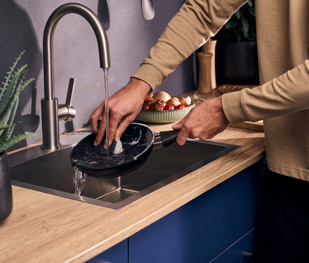 Ein Mann wäscht eine schwarze Bratpfanne aus dem Henckels by ZWILLING »Breeze« Bratpfannen-Set mit einem Schwamm unter fließendem Wasser in einem Küchenwaschbecken.