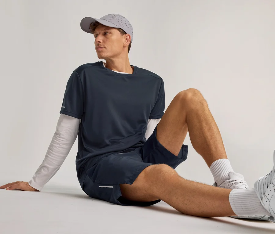 Mann sitzt am Boden, trägt eine graue Kappe, ein marineblaues T-Shirt über einem weißen Langarmshirt und marineblaue Sportshorts, blickt nach rechts.