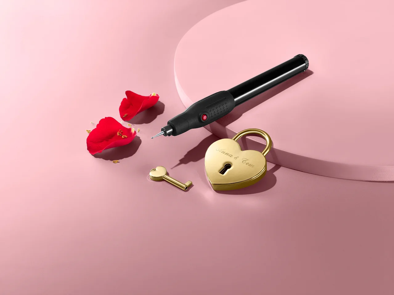 Liebesschloss mit Gravierstift, Schlüssel und Rosenblättern auf rosa Hintergrund.