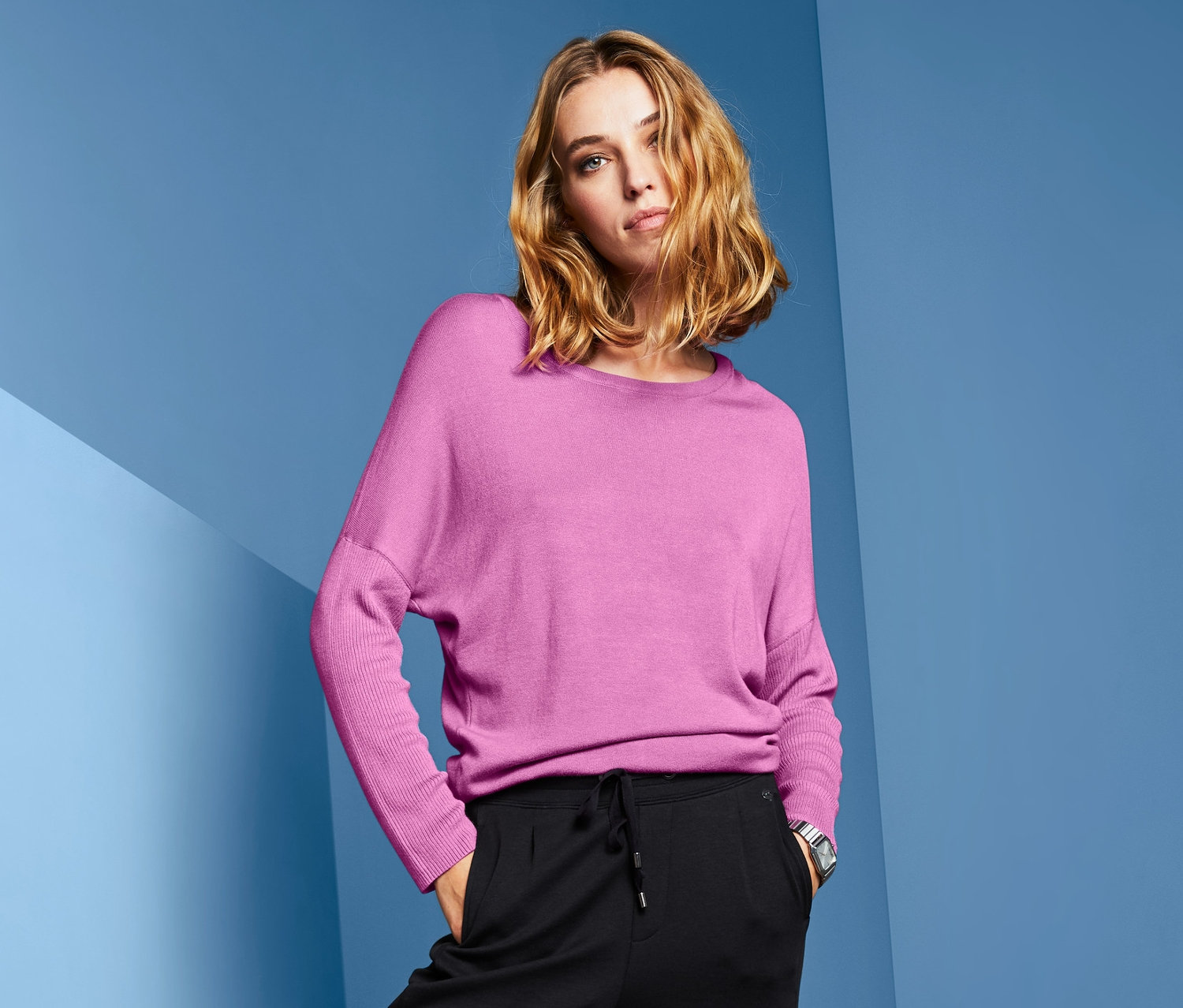 Frau posiert in Feinstrickpullover mit Fledermausärmeln in Pink, schwarzer Hose und silbernen Schuhen.