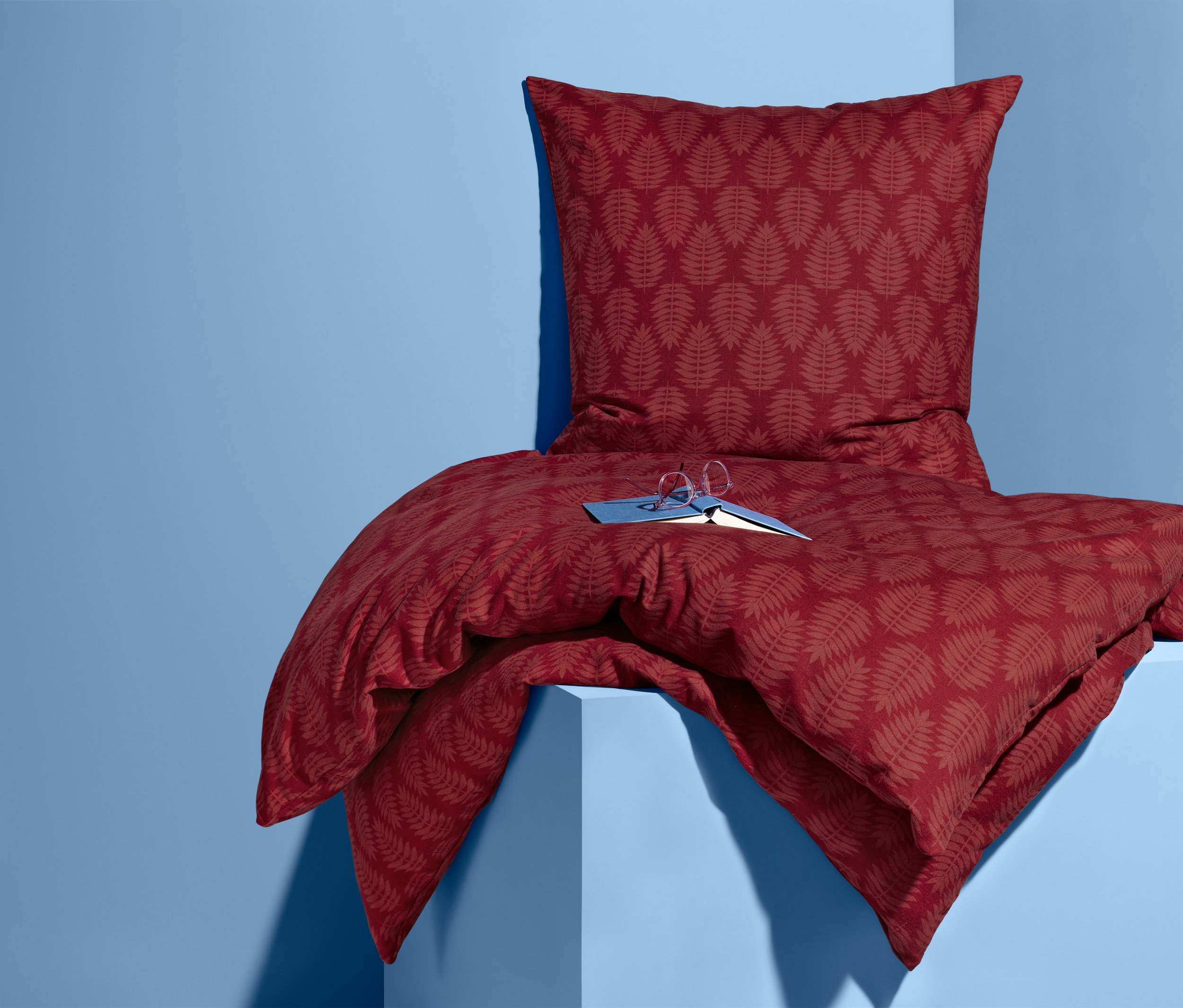 Ein Set Biber-Bettwäsche mit Blattmuster in Rot liegt auf einem blauen Sockel, darauf liegen ein Buch und eine Brille.
