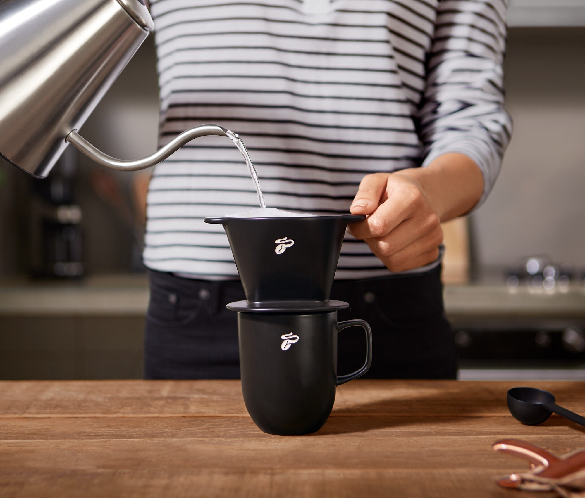 Person gießt Wasser aus einem Kessel in einen schwarzen Kaffeehandfilter Gr. 101 mit Bohnenmotiv, der auf einer Tasse steht.