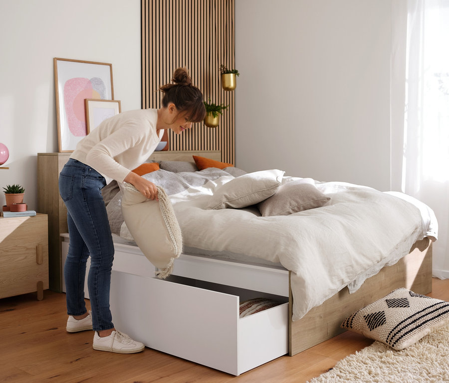 Eine Frau legt ein Kissen in das Staufach des Stauraumbetts. Im Hintergrund sind 4 Ordnungsboxen und der Teddysessel »Jack« zu sehen.