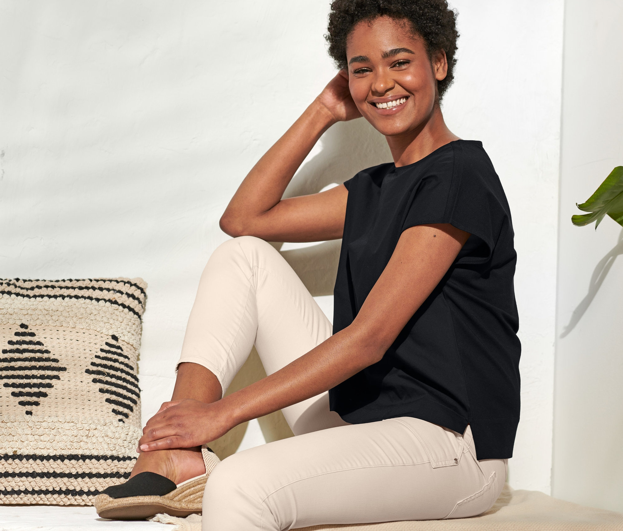 Eine lächelnde Frau sitzt, trägt ein schwarzes Blusenshirt im Materialmix und helle Hosen.