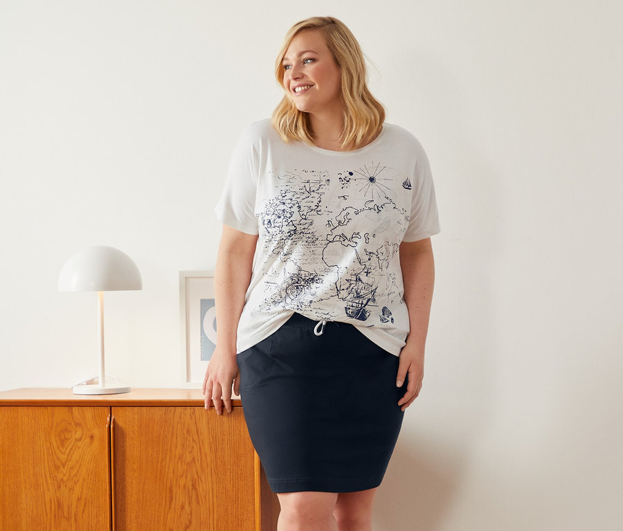 Eine Frau steht neben einem Schrank und trägt ein weißes Shirt mit Rundhalsausschnitt und einen Sweatrock.