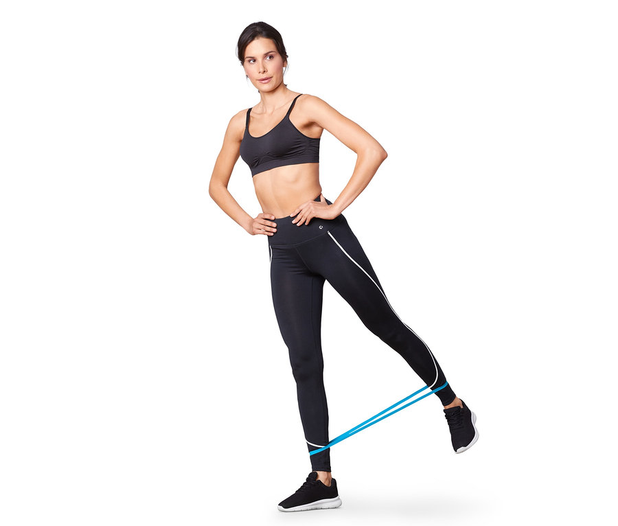 Frau trainiert mit einem blauen Fitnessband, sie trägt einen schwarzen Sport-BH und Leggings. Im Hintergrund ist ein weißer Hintergrund. Sie hat die Hände in den Hüften.