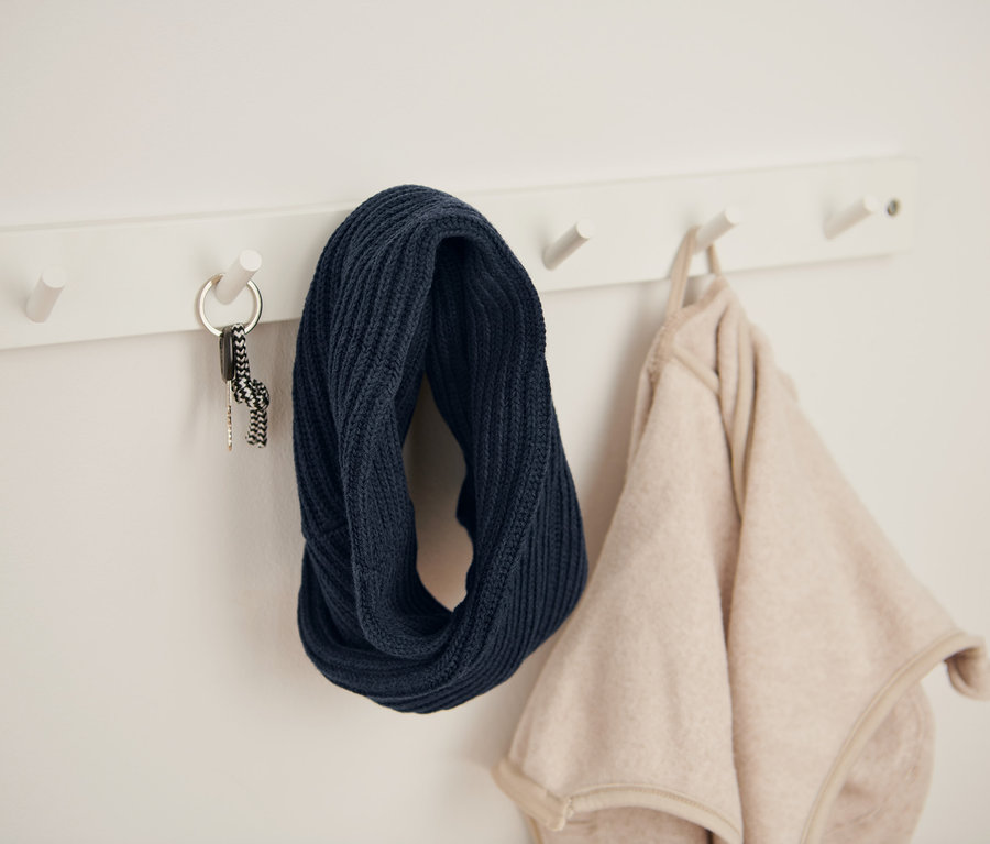 An einer weißen Wandgarderobe hängen ein dunkelblauer Strickloop, Schlüssel und eine beige Jacke.