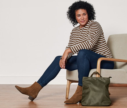 Eine Frau sitzt in einem Sessel und trägt ein gestreiftes Langarmshirt, Jeggings und Stiefeletten aus Veloursleder. Neben dem Sessel steht ein Rucksack.