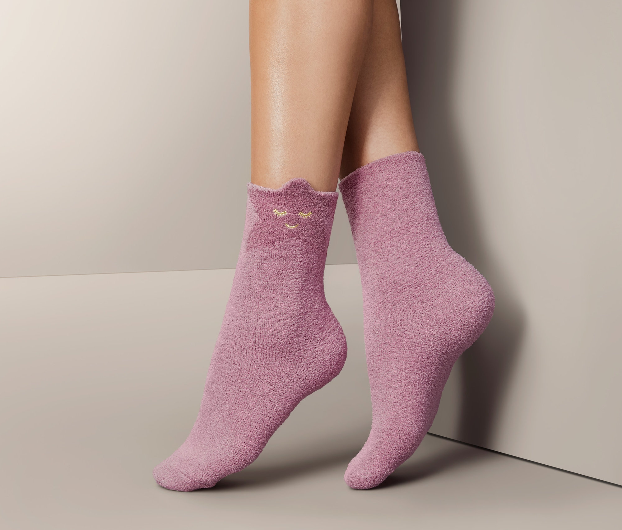 Beine in einem Paar flauschiger, rosa Socken mit Gesichtsmotiv.