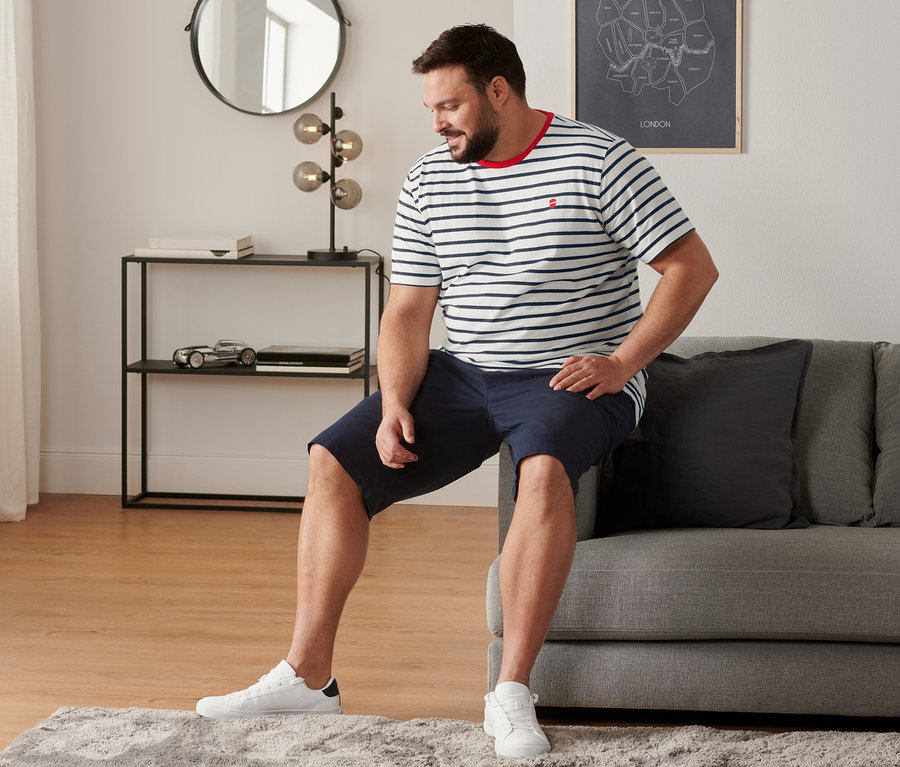 Mann sitzt auf Sofa mit Sneaker, Chino-Shorts, navy, und 2 T-Shirts mit Rundhalsausschnitt, gestreift.