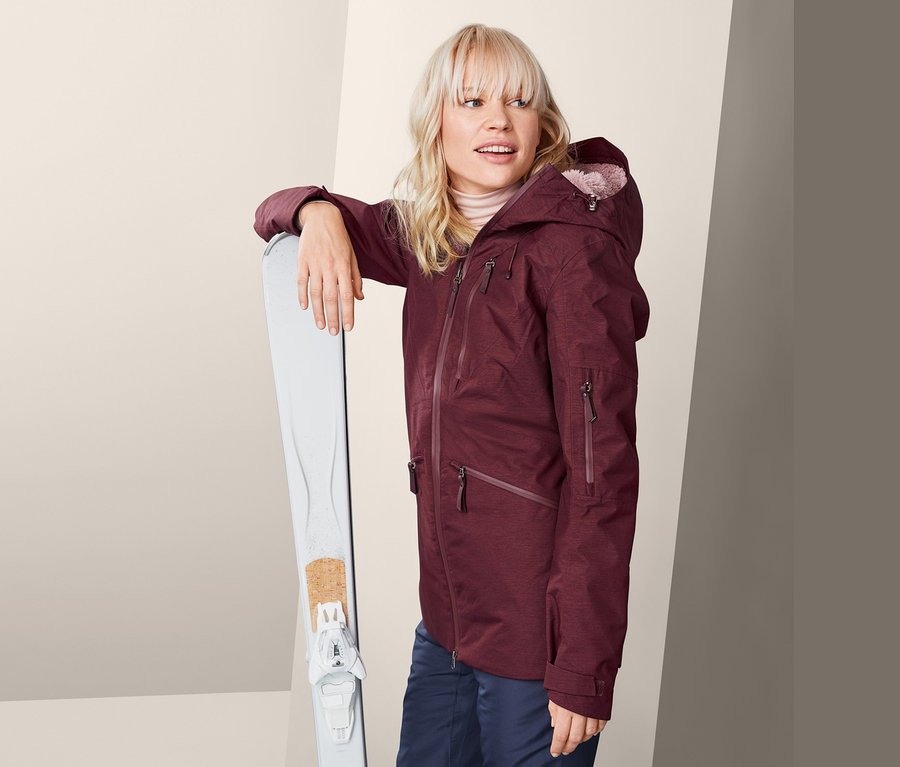 Blonde Frau in bordeauxfarbener Ski- und Snowboardjacke steht neben weißen Skiern.