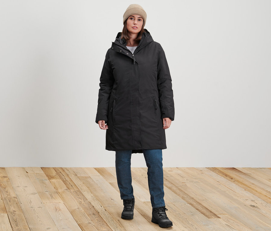 Eine Frau steht und trägt einen schwarzen Thermomantel, eine dunkelblaue Slim-Denim »Fit Emma« und eine sandfarbene Strickmütze.