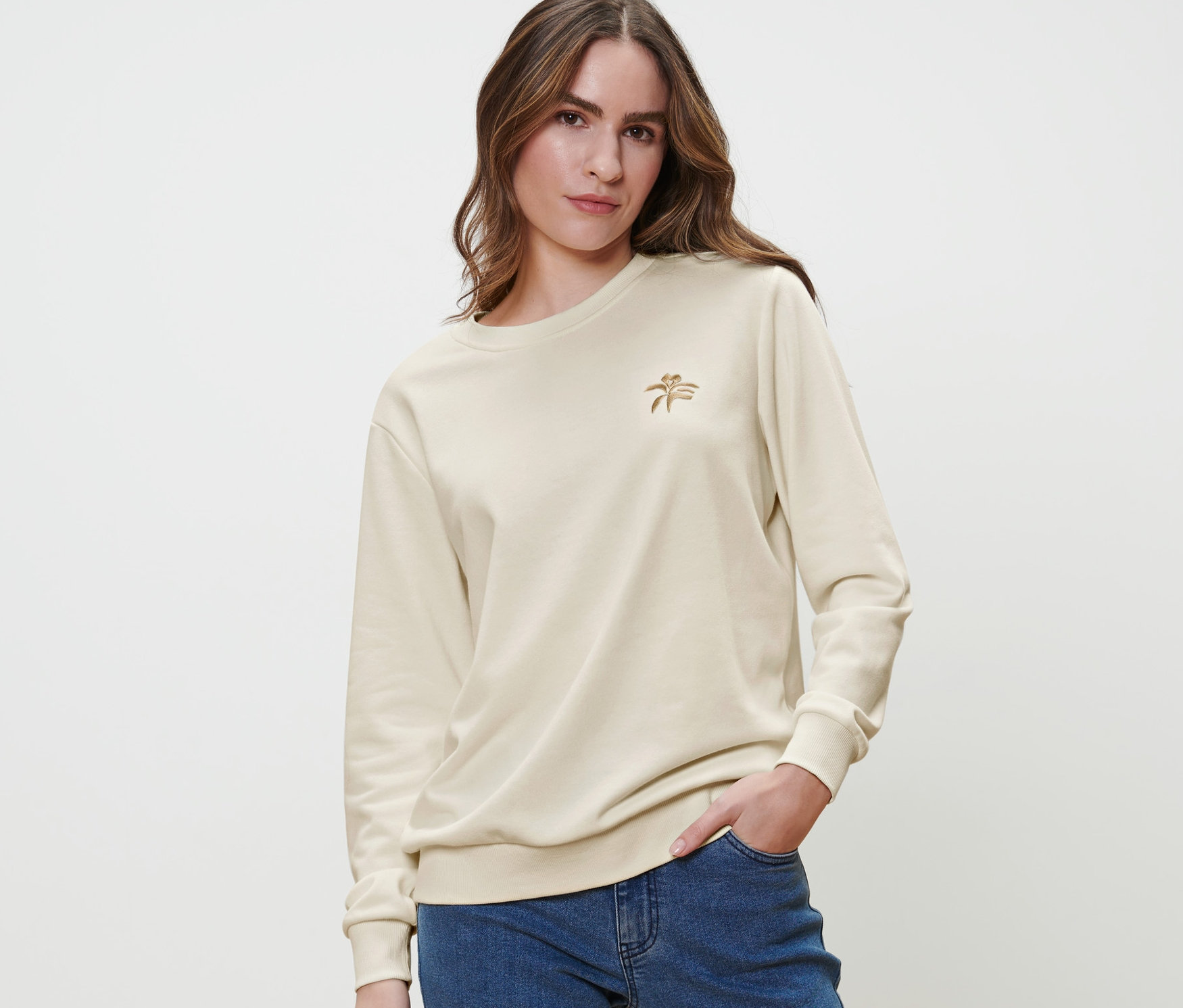 Frau posiert in einem cremefarbenen Sweatshirt mit Stickerei und blauen Jeans.