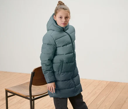 Mädchen trägt Kinder-Thermomantel mit Farbverlauf und steht neben einem Holzstuhl.