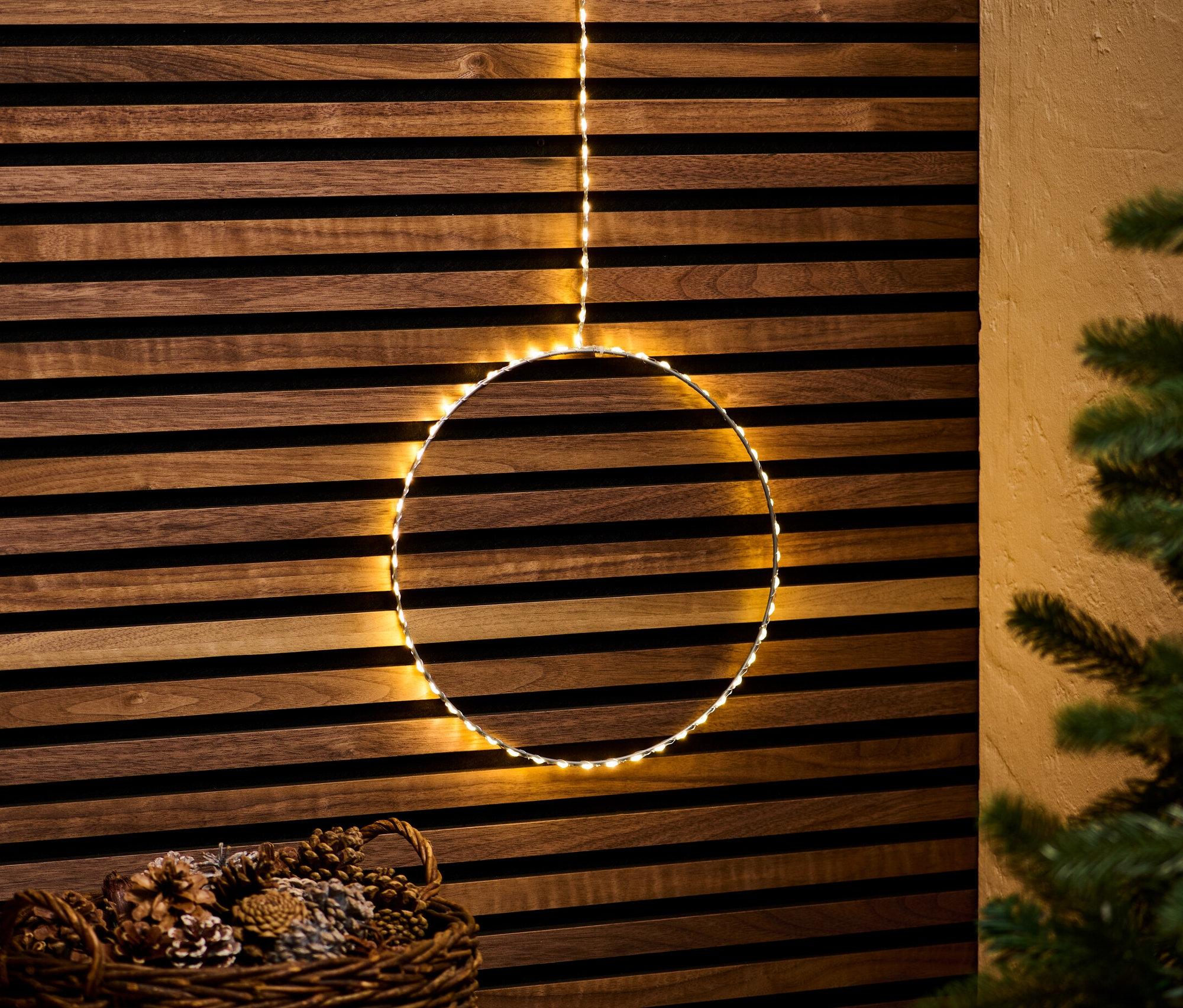 Ein hängender Outdoor-LED-Kreis vor einer Holzwand, darunter ein Korb mit Tannenzapfen.