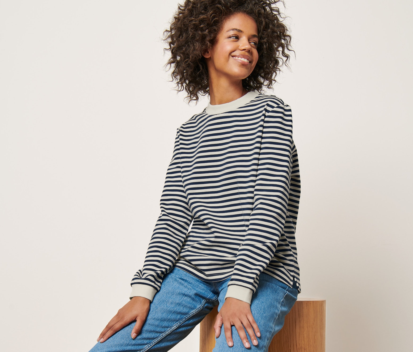 Frau sitzt auf einem Holzhocker und trägt ein gestreiftes Sweatshirt.