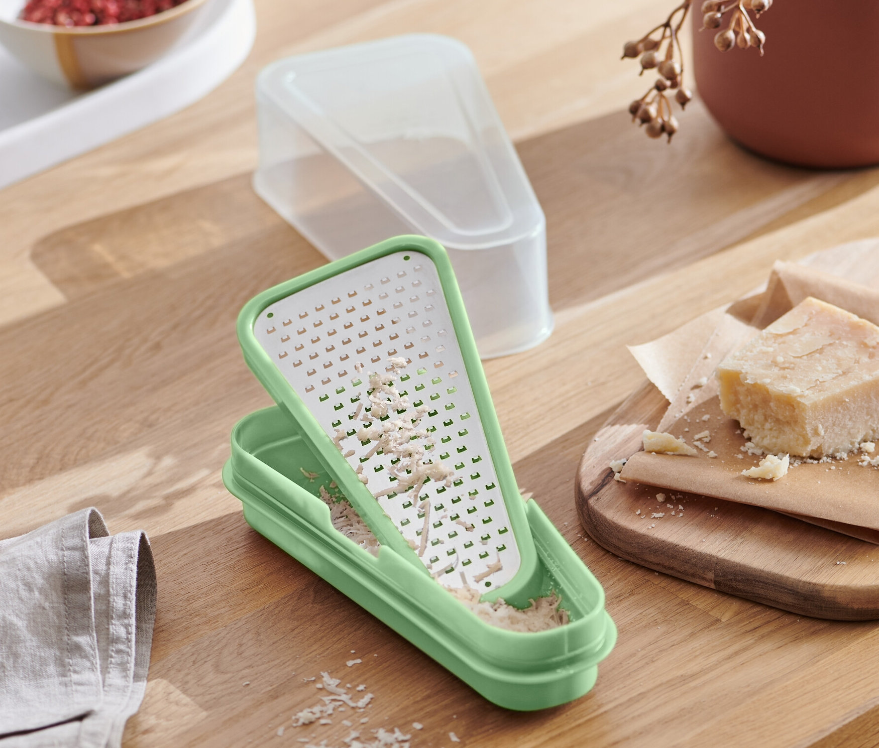 Eine grüne 2-in-1-Parmesanreibe mit geriebenem Käse steht auf einem Holztisch neben einem Teller Pasta.