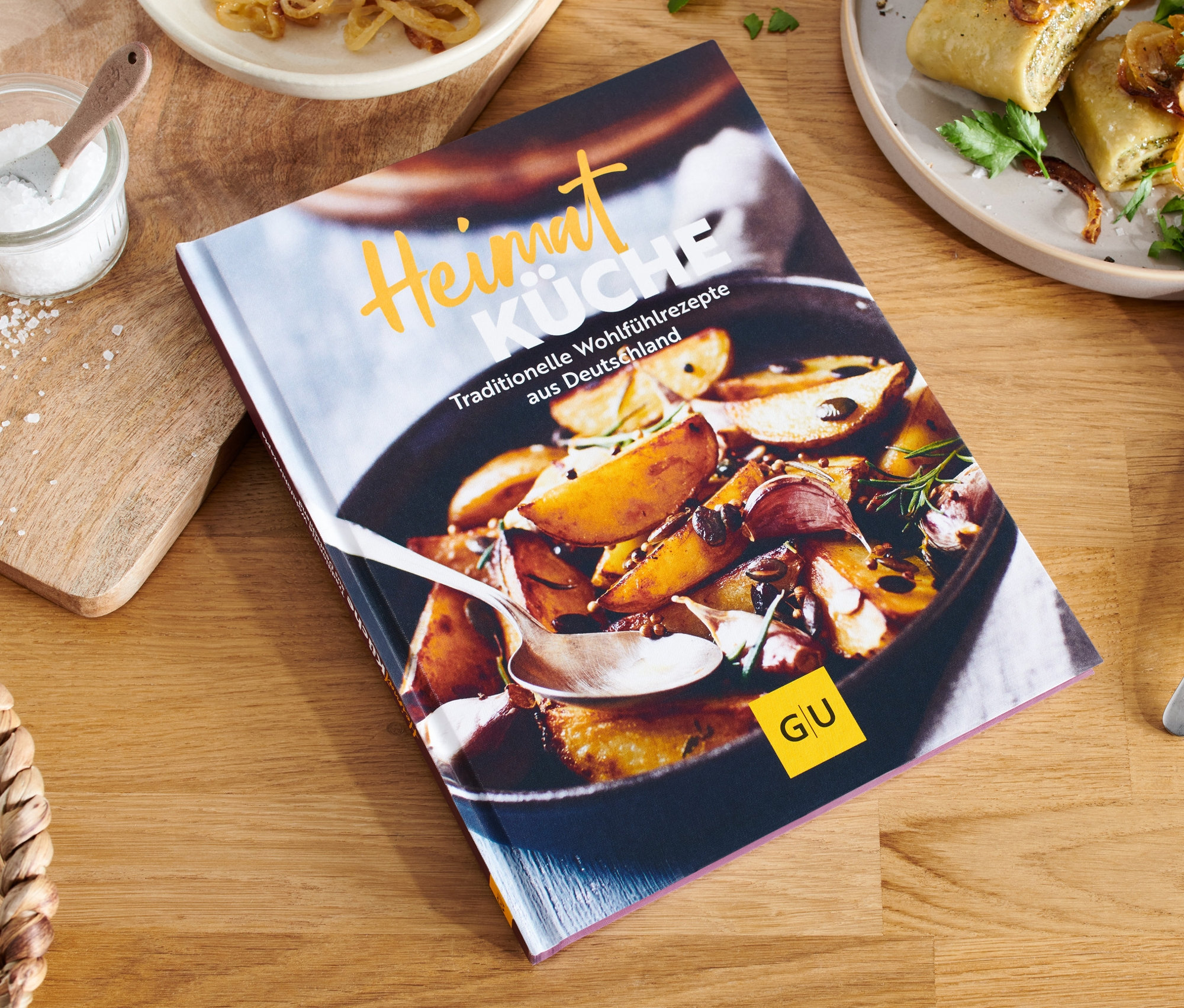 Auf einem Holztisch liegt das Buch "Heimatküche", daneben ein Teller mit Speisen und ein Salzlöffel.