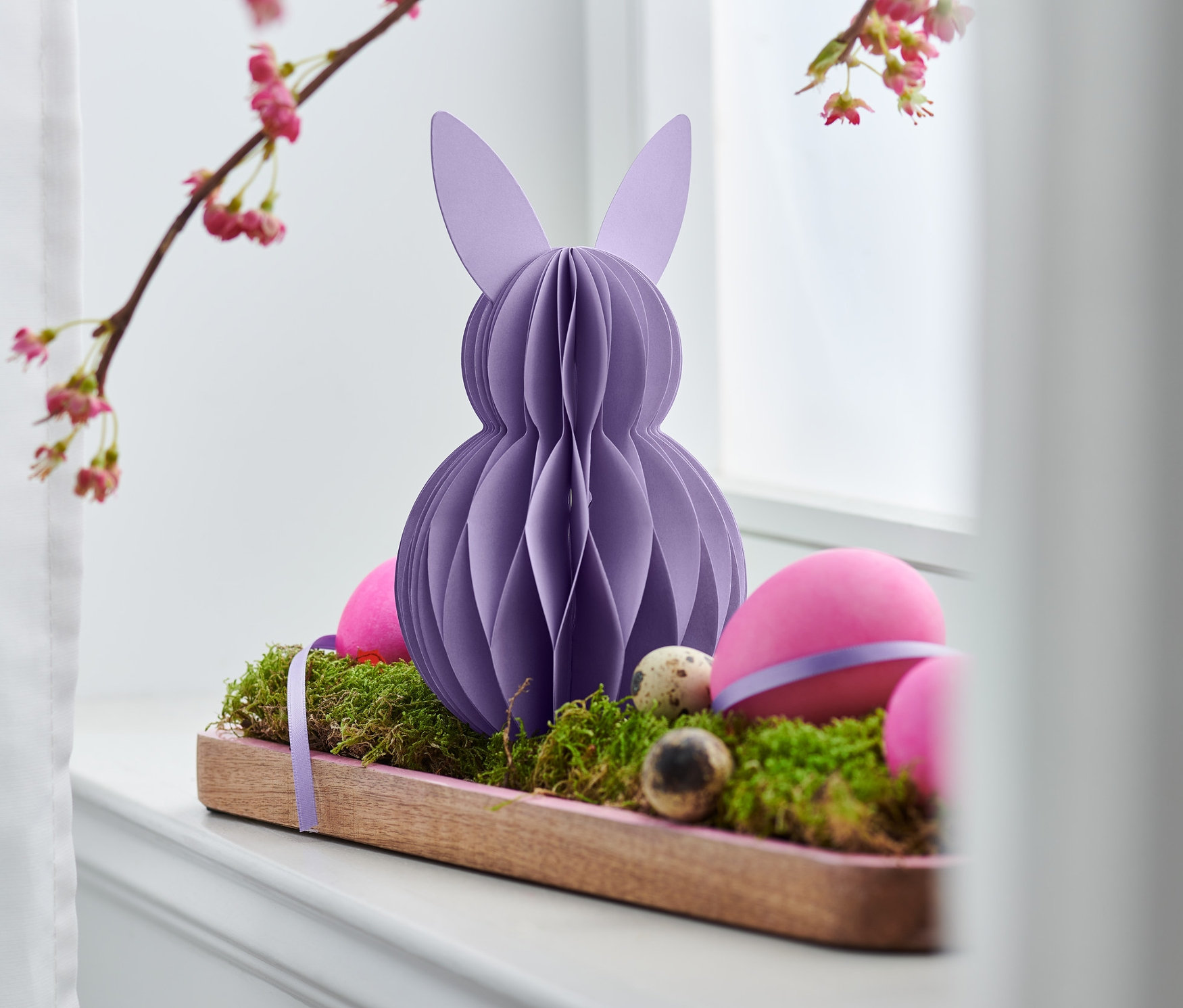 Ein lila Papier-Osterhase steht auf einem Holztablett mit Moos und Eiern.