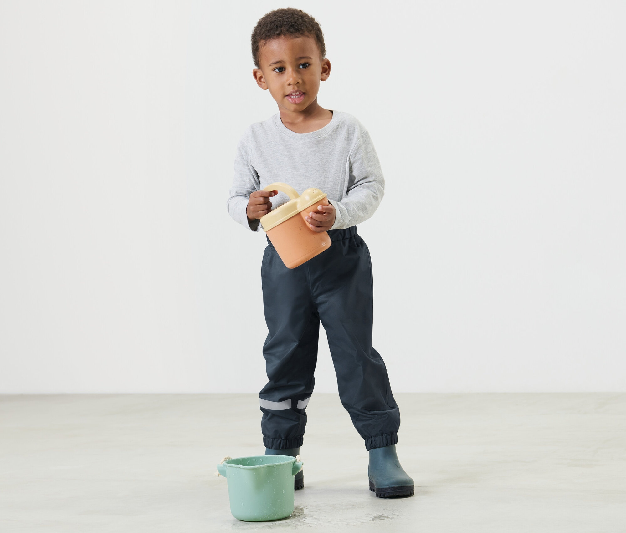 Junge hält einen orangefarbenen Eimer und trägt eine dunkelblaue Kinder-Regenhose.