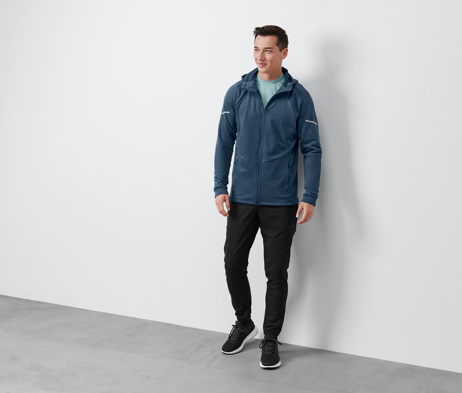 Ein Mann steht und trägt eine blaue Thermojacke, eine schwarze Thermojacke, eine Thermo-Funktionshose und schwarze Schuhe.