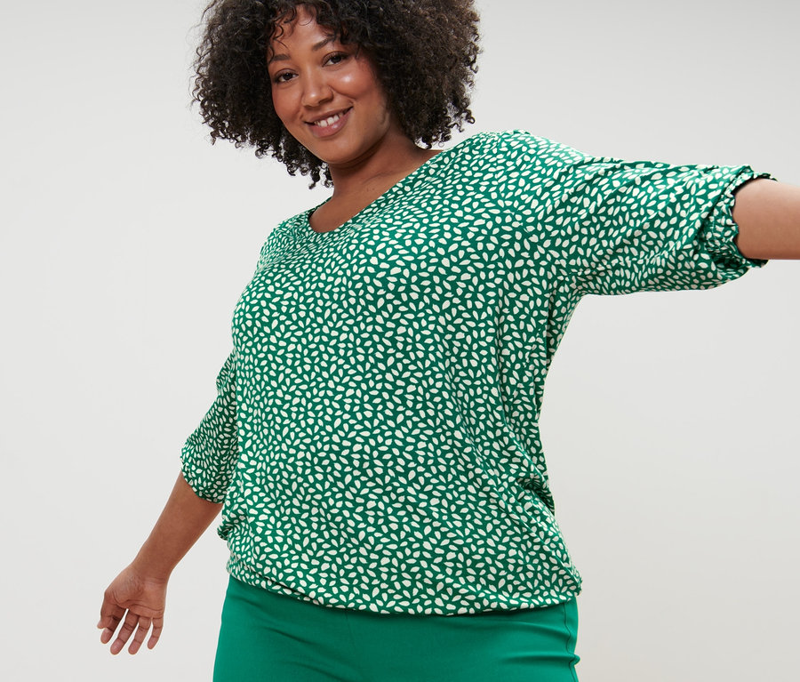 Frau posiert in grünem, bedrucktem Blusenshirt und grüner Stretchhose.