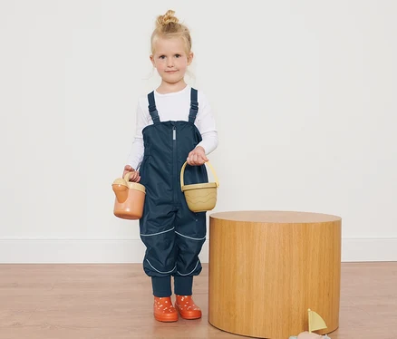 Ein Mädchen in Kleinkinder-Regenhose hält Spielzeug neben einem Holztisch.