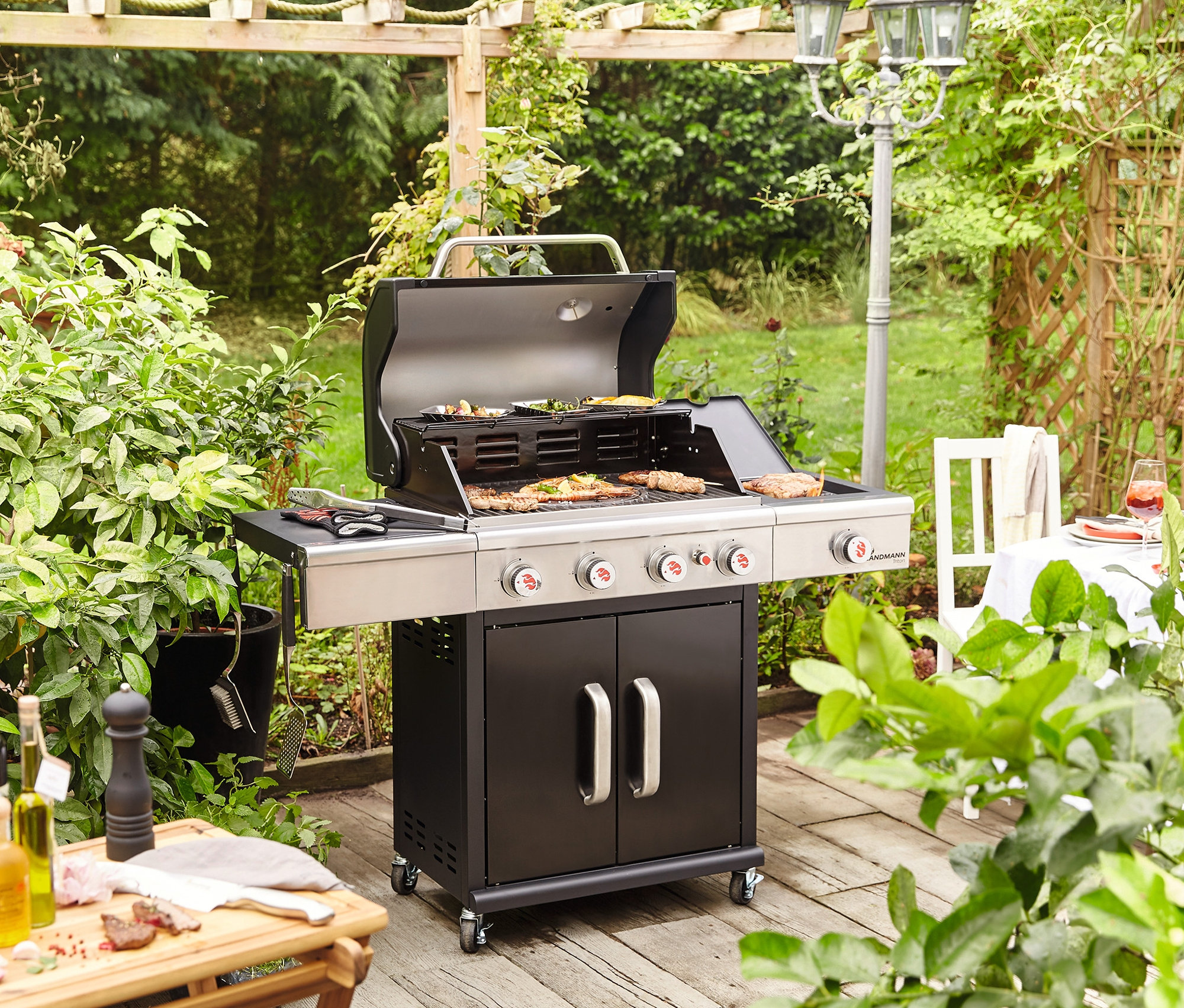 Auf einer Holzterrasse steht ein Landmann Gasgrill »Triton maxX BR 4.1« mit geöffnetem Deckel und Essen auf dem Rost.