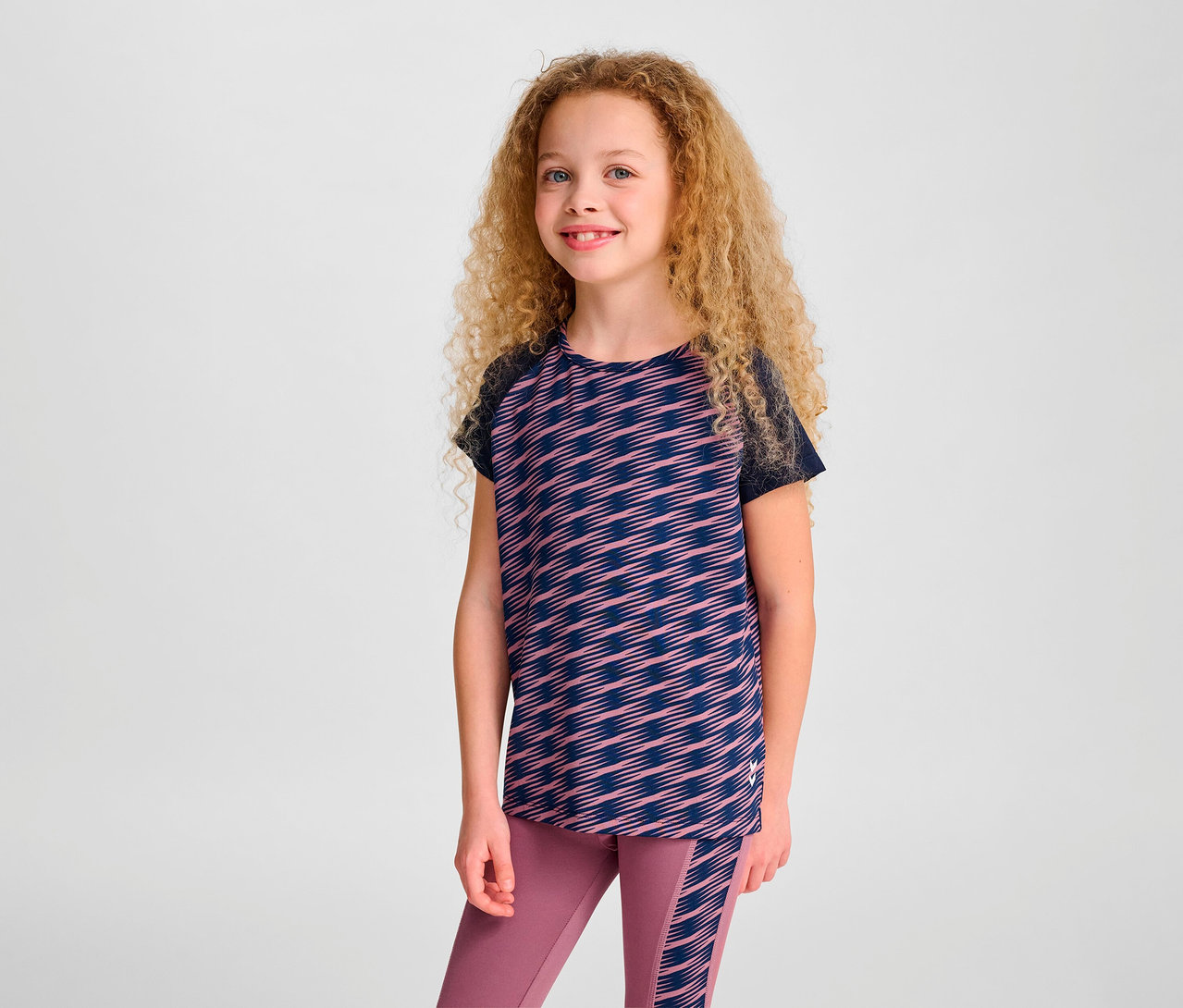 Mädchen mit lockigen Haaren posiert in einem HUMMEL HMLJR Mix T-Shirt S/S und Leggings.