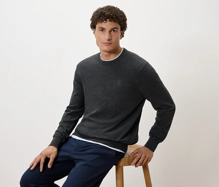 Mann sitzt auf einem Hocker, trägt einen grauen Pullover und dunkelblaue Hosen.