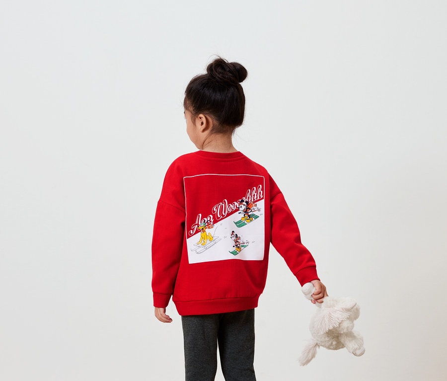 Mädchen mit dem Rücken zur Kamera trägt ein rotes Sweatshirt mit einem Bild auf dem Rücken und hält ein Plüschtier in der Hand.