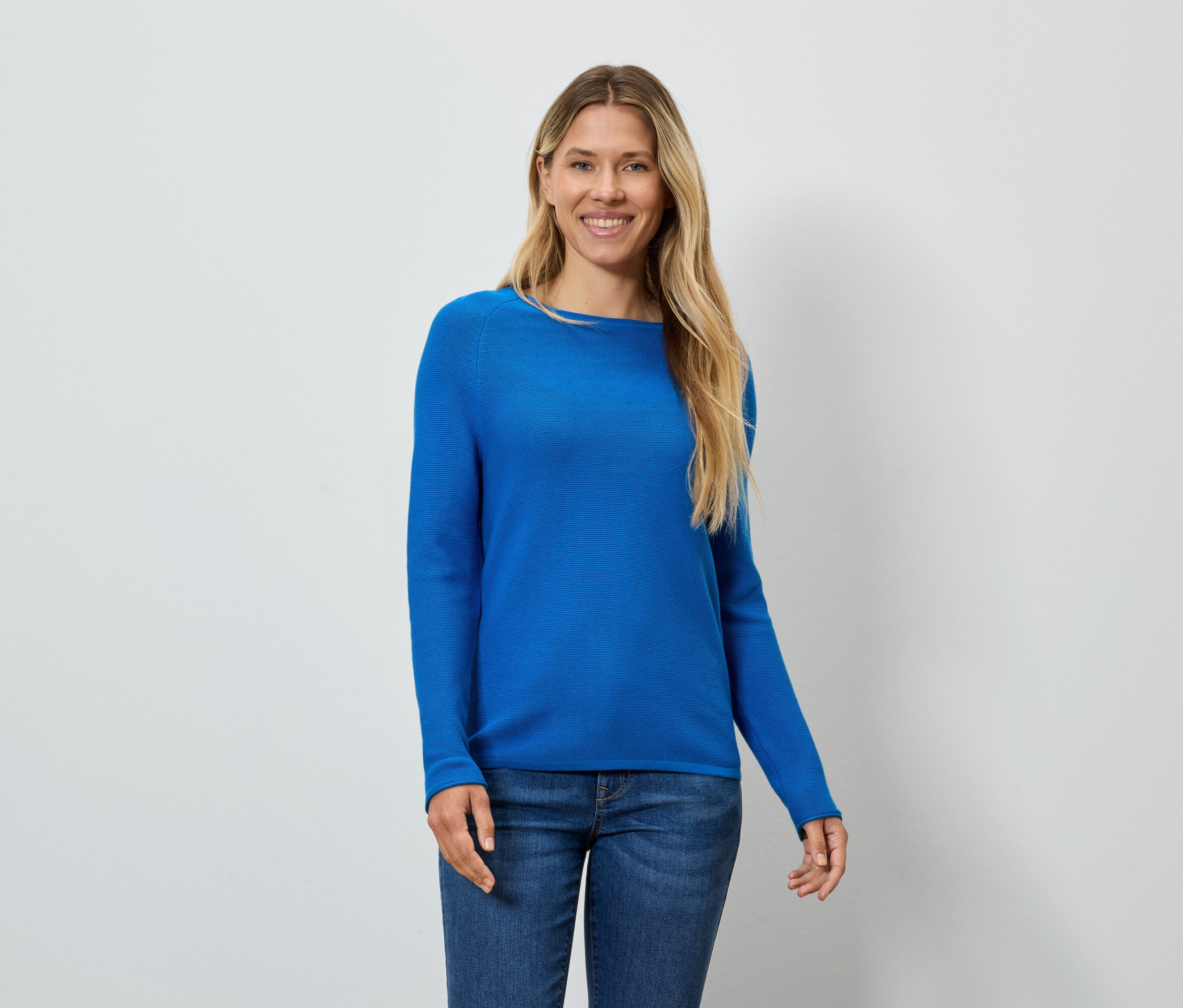 Frau mit blonden Haaren, blauem Pullover und Jeans lächelt.