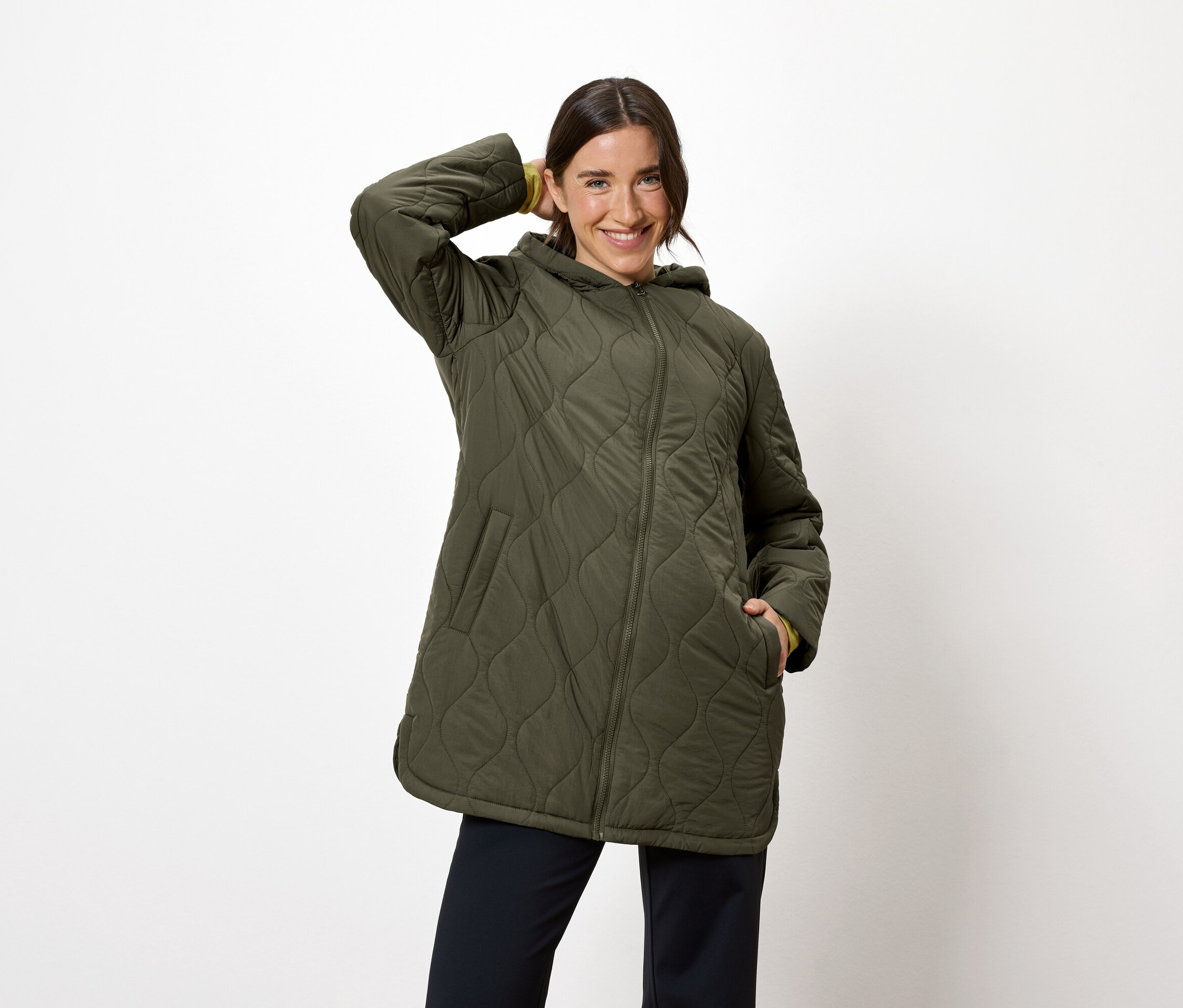 Frau posiert in olivgrüner Steppjacke mit Kapuze und marineblauer Hose.