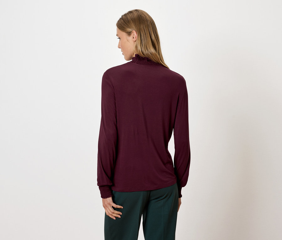 Frau trägt ein bordeauxrotes Langarm-Rollkragenshirt und grüne Hosen, von hinten gesehen.