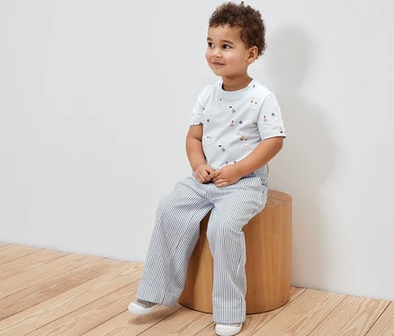 Ein Junge sitzt auf einem Hocker und trägt eine Kinder-Webhose und ein T-Shirt mit Aufdruck.