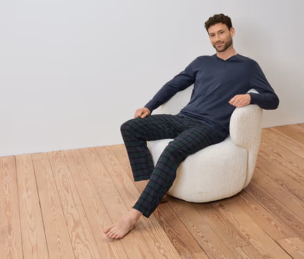 Mann sitzt in einem Sessel mit einem blauen Langarmshirt und einer karierten Hose.