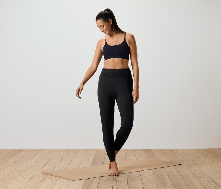 Frau steht auf einer Yogamatte und trägt einen schwarzen Sport-BH und schwarze Leggings.