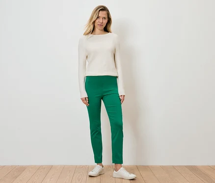 Eine Frau steht und trägt einen cremefarbenen Pullover, grüne Hosen und weiße Turnschuhe.