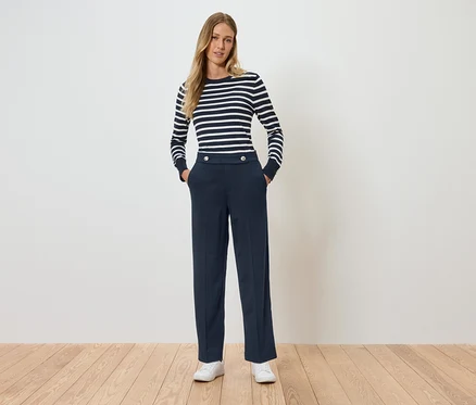 Eine Frau steht mit den Händen in den Hosentaschen. Sie trägt ein gestreiftes Langarmshirt und dunkelblaue Hosen.