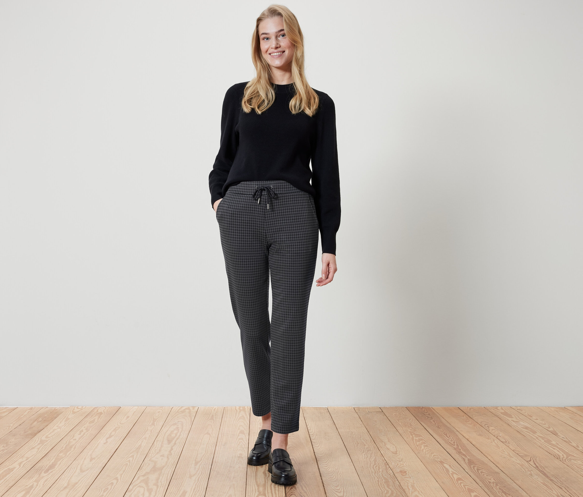 Eine Frau steht mit den Händen in den Taschen einer karierten Hose. Sie trägt einen schwarzen Pullover und schwarze Schuhe.